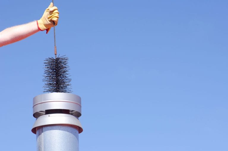 Gloved hand lowering a chimney sweep brush into a metal chimney flue cap against a clear blue sky
