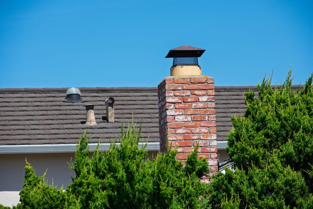 Brick chimney with a black metal cap against a clear blue sky, surrounded by trees.