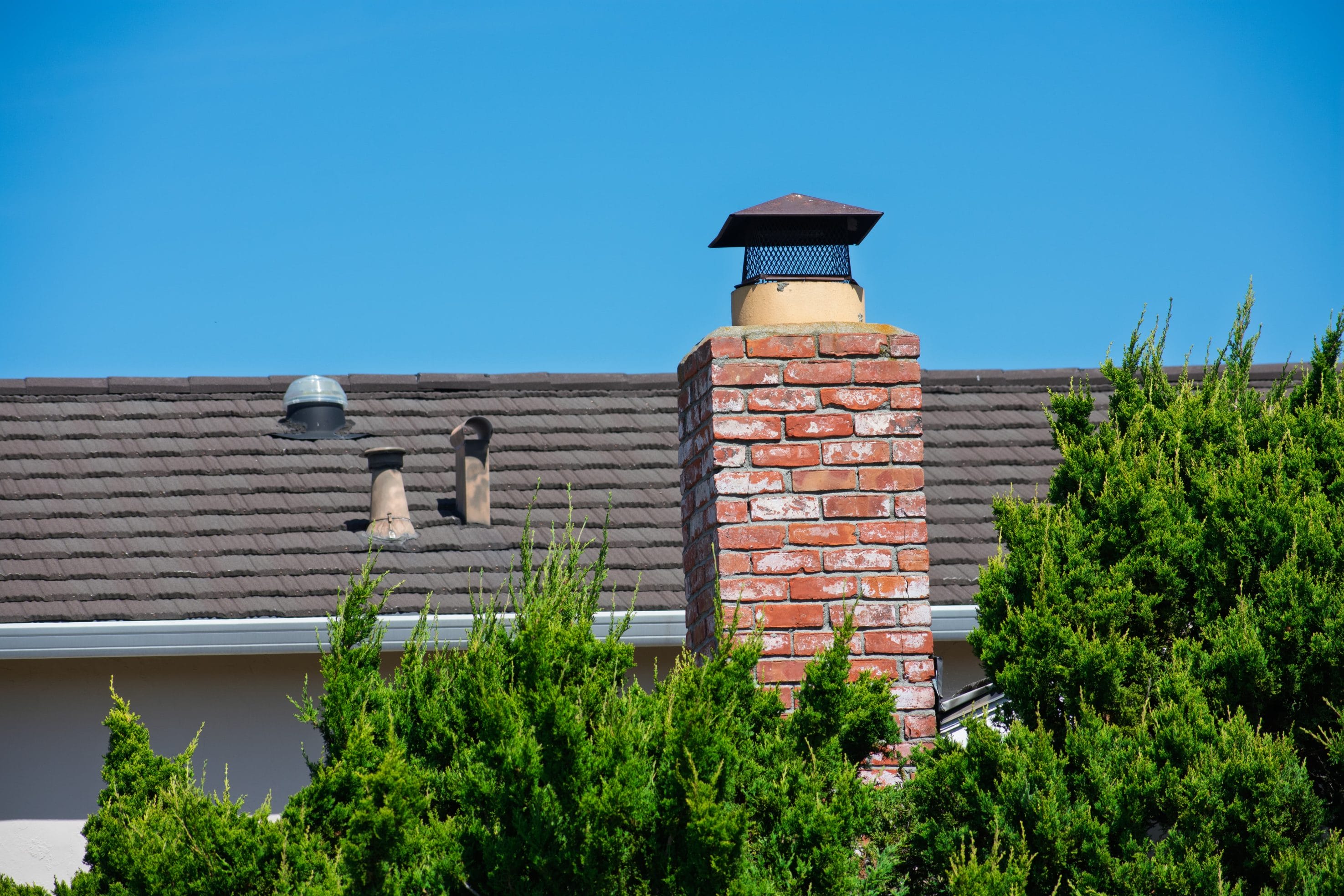 Brick chimney with a black metal cap against a clear blue sky, surrounded by trees.